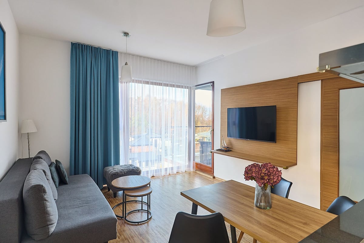 Ett modernt vardagsrum med en soffa, ett matbord, en tv och ett stort fönster med gardiner.