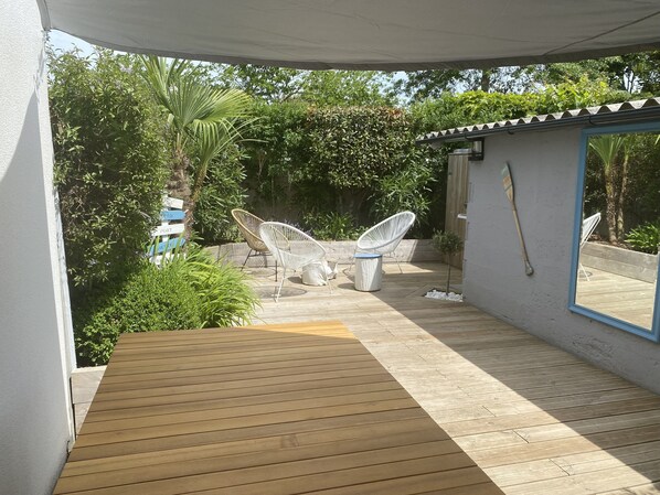 A covered patio area with a wooden deck, two white wicker chairs, a small table, and a trash bin. - Very pleasant house close to the beach and shops (Le Pouliguen)