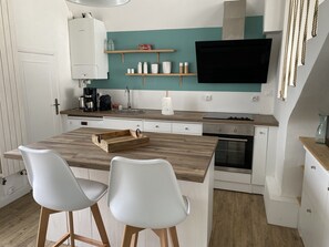 A compact kitchen with a wooden island, two white chairs, a TV mounted above the island, and a teal accent wall. - Very pleasant house close to the beach and shops (Le Pouliguen)