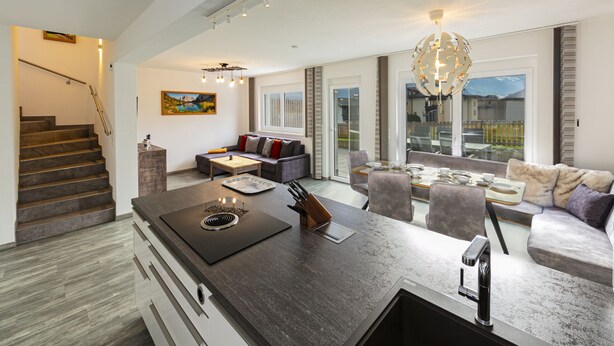 A modern kitchen with a central island, a dining area with a table set for four, a sofa, and a staircase leading to an upper level.