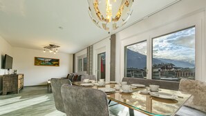 A modern dining area with a glass table, grey upholstered chairs, and a view of mountains through large windows.