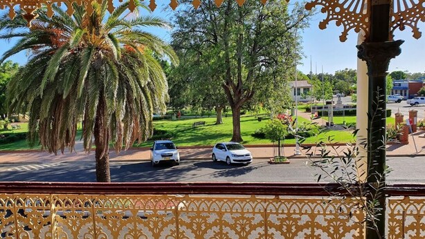 Une vue depuis un balcon orné de balustrades en métal ouvragé, avec un palmier et deux voitures garées dans une rue, un parc se dessinant à l’arrière-plan.