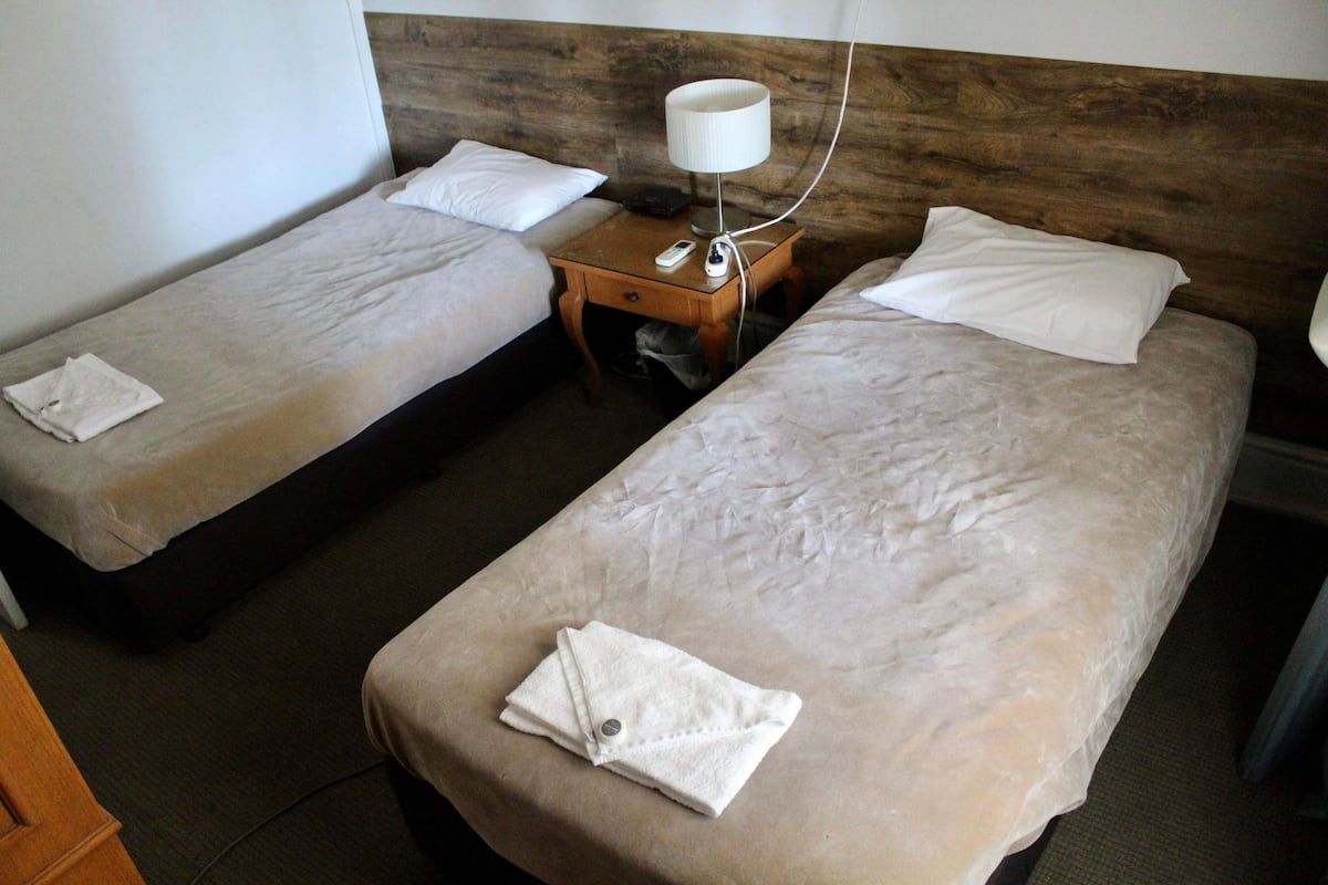 Habitación de hotel con dos camas individuales, una mesita de noche de madera con lámpara y una pequeña mesa con un control remoto.