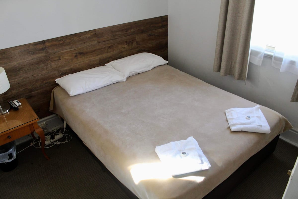 Una cama bien hecha con cabecera de madera, una mesita de noche con lámpara y un cubo de basura pequeño.