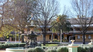 Une fontaine ornée d’une statue au centre, entourée de haies et d’arbres bien entretenus. Au fond, on aperçoit un bâtiment sur lequel est apposée une enseigne indiquant « Hôtel ».