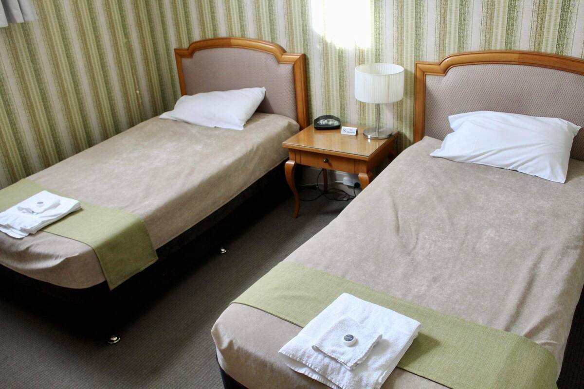 Una habitación de hotel con dos camas individuales, cada una con una almohada, una mesita de noche con una lámpara y un pequeño atril.