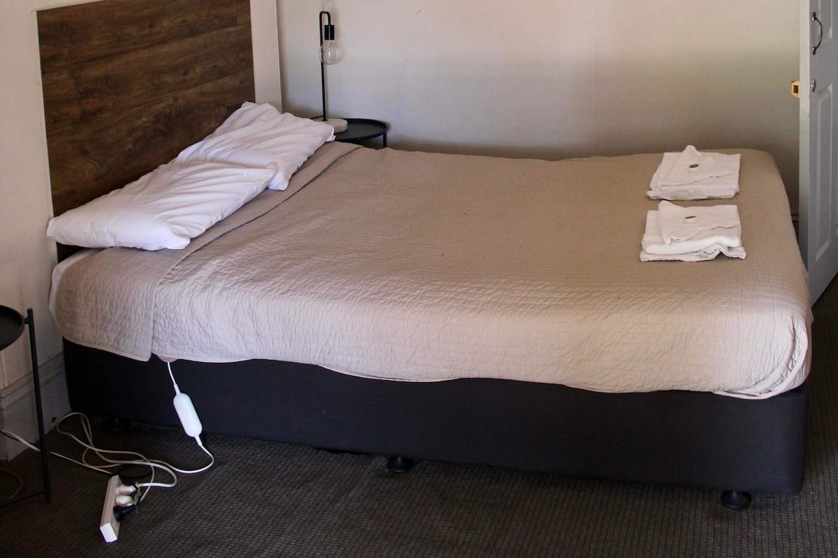 Una cama bien hecha con cabecera de madera, un cuadro enmarcado en la pared y un enchufe a un lado.