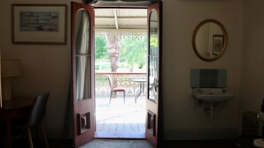 Une porte ouverte donnant sur un balcon avec une chaise et une table, un tableau encadré au mur, un miroir rond et un lavabo avec un pare-éclaboussures carrelé.