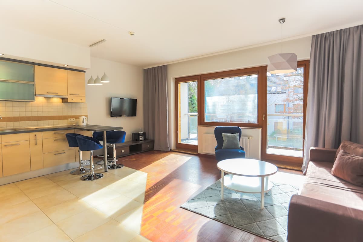 Un apartamento moderno con cocina, comedor y sala de estar. Cuenta con un amplio ventanal corredera al exterior, un televisor de pantalla plana montado en la pared y una zona de estar acogedora con sofá y sillones.