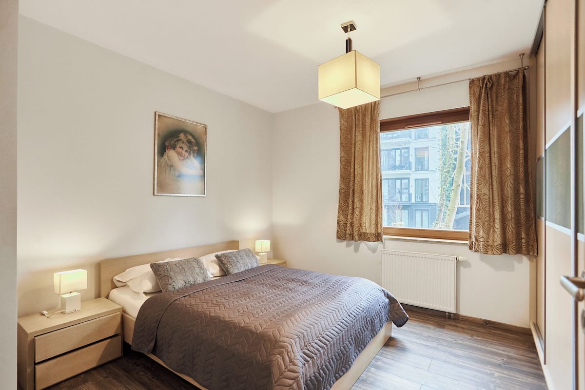 Un dormitorio con cama, mesitas de noche, lámpara colgante, ventana con cortinas y radiador.