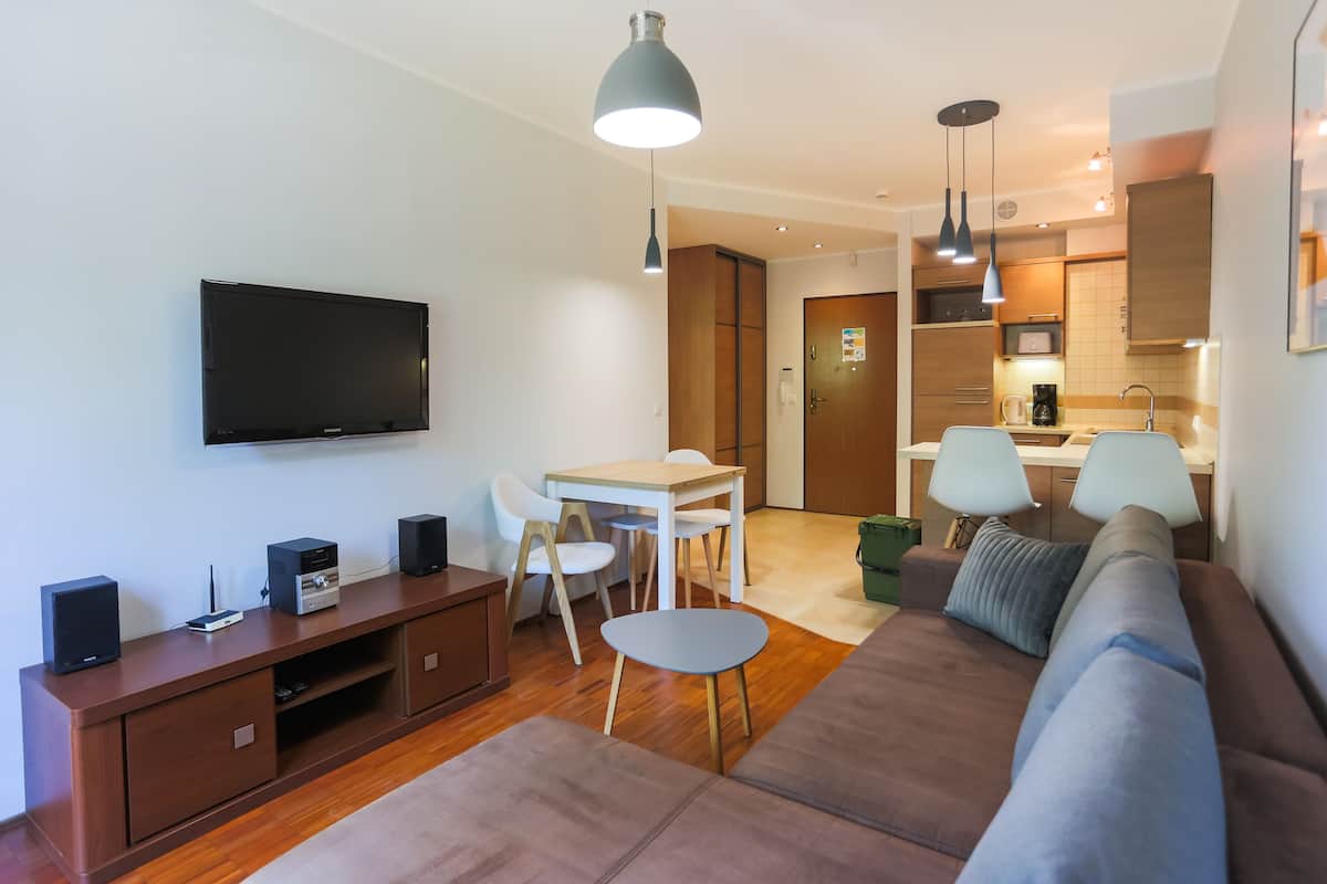 Una sala de estar moderna con televisor de pantalla plana, mueble de madera para entretenimiento, sofá marrón, una mesita redonda y un comedor con mesa y sillas blancas.
