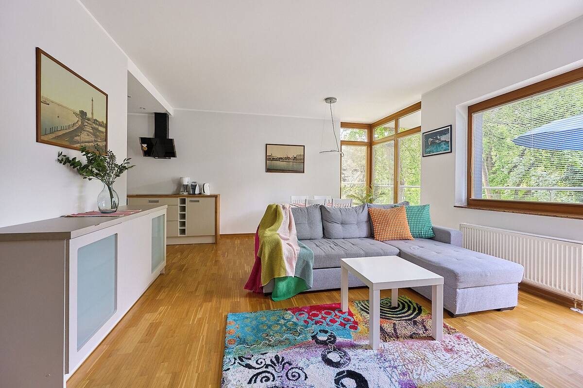 Una sala de estar moderna con un sofá gris, una mesa de centro blanca, un tapete estampado y vistas amplias a través de grandes ventanales.