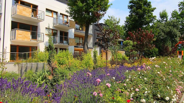 Un moderno edificio de apartamentos con balcones, rodeado por un jardín con diversas flores y plantas.