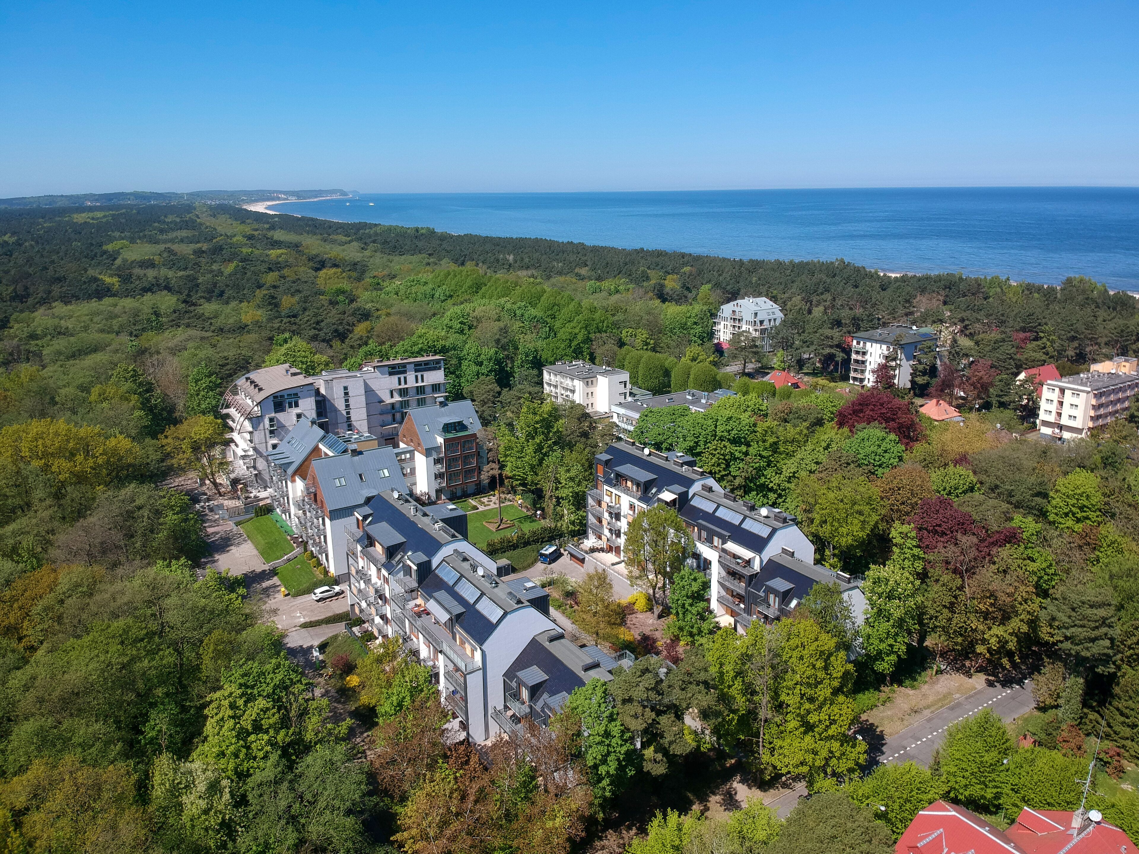 An aerial view of a residential area with modern buildings, surrounded by dense greenery and a coastline in the background.