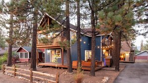 A two-story log cabin with a deck, surrounded by pine trees.