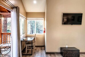A room with a wooden dining table, chairs, a television mounted on the wall, and a window with a view of trees. - Main House + Guest House · Spa · Game Room (Big Bear)