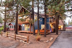 A two-story cabin with a deck, surrounded by pine trees. - Main House + Guest House · Spa · Game Room (Big Bear)