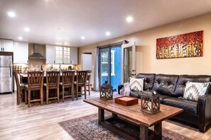A modern living room with a dark leather sofa, wooden dining table, and chairs. A kitchen with white cabinets and a refrigerator is visible in the background. - Main House + Guest House · Spa · Game Room (Big Bear)