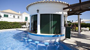 A pool area with a covered seating area, a white building with green shutters, and a clear blue sky.