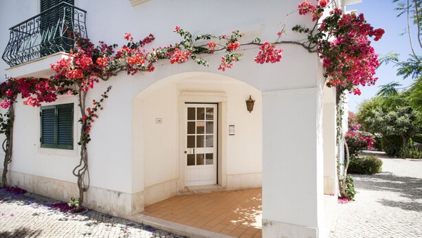 A white building with a white door, a small balcony, and pink flowers growing over the entrance.