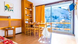 Ein Hotelzimmer mit einem hölzernen Bett, einem Esstisch mit Stühlen, einem Bücherregal, einem großen Fenster mit Blick auf einen verschneiten Berg und einem Gemälde an der Wand.