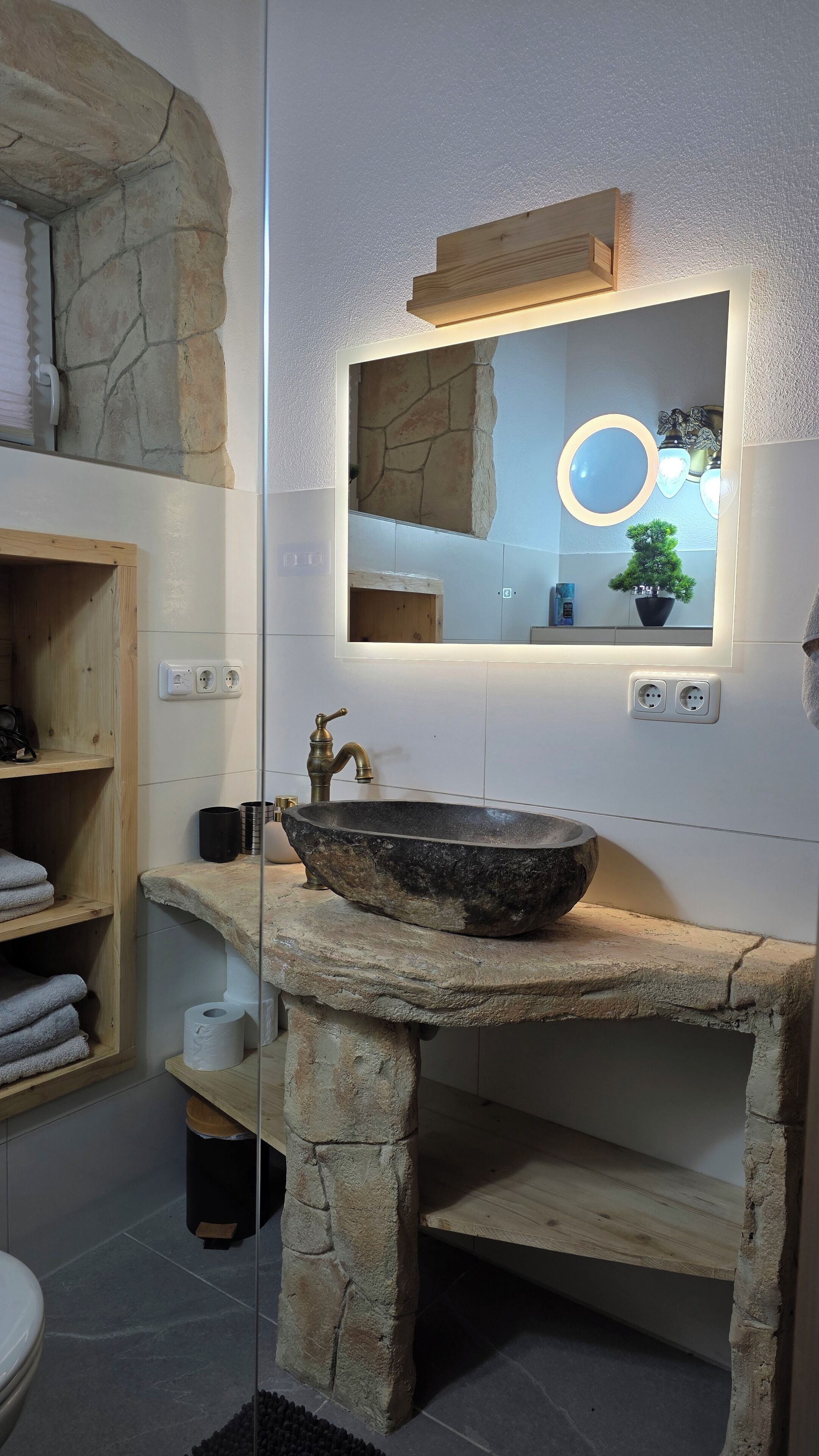 Ein Badezimmer mit einem Steintonne, Holzregalen, einem Spiegel mit runder Beleuchtung und einer Steinwand.