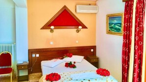 A hotel room with a bed, a red and white triangular wall decoration, an air conditioner, a red chair, and a window with red curtains.