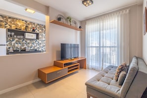 A modern living room with a flat-screen TV, built-in shelving, a sofa, and a large window with sheer curtains. - 2 Bedroom Apartment with swimming pool, gym, laundry and hammocks on the windows (São Paulo)