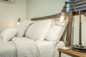Una cama bien hecha con edredón blanco y almohadas, una mesita de noche con una lámpara y un pequeño objeto decorativo sobre la mesa.
