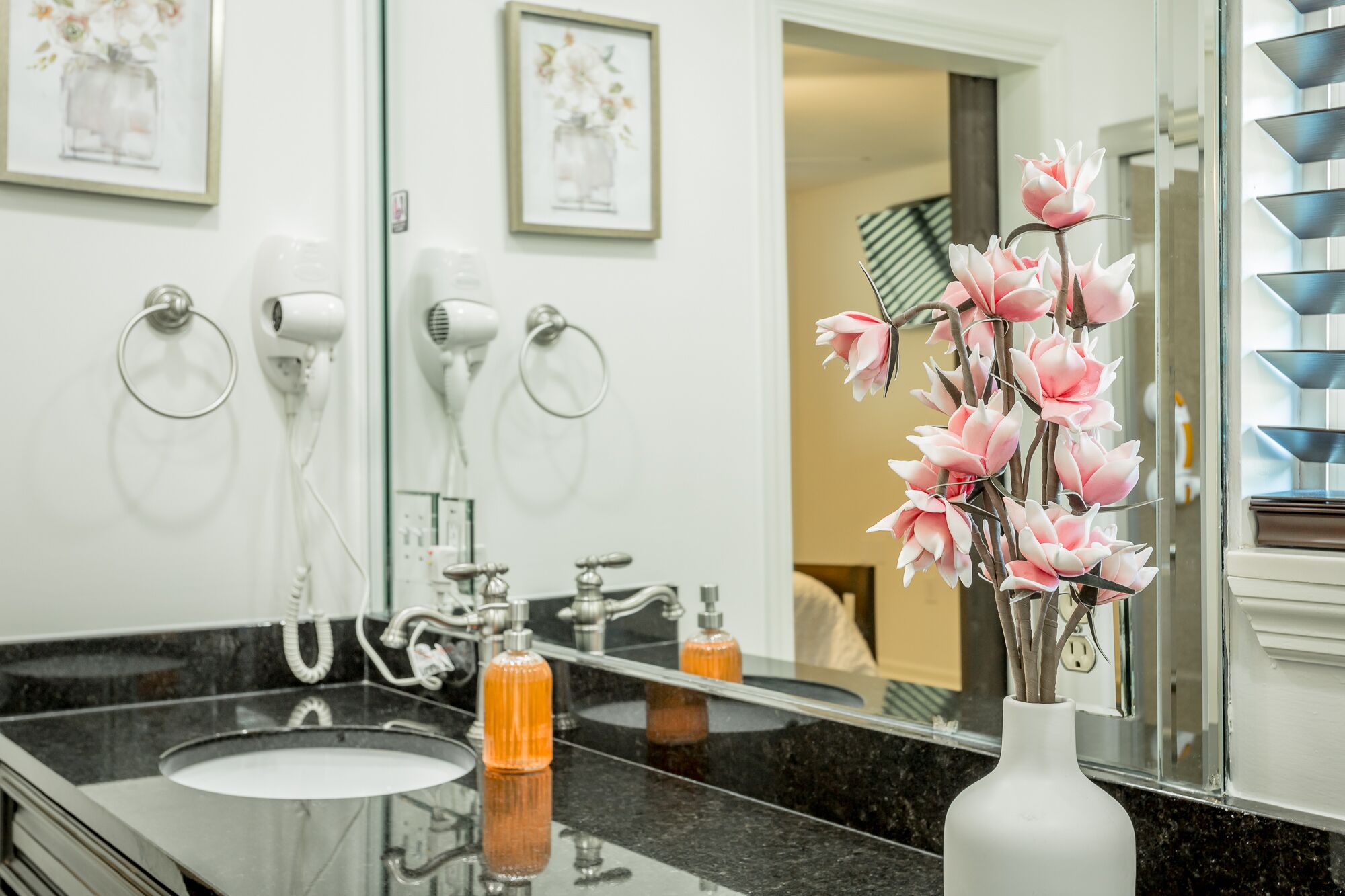 Un baño con lavamanos de mármol negro, dos secadores de pelo, un espejo y un florero con flores rosas.