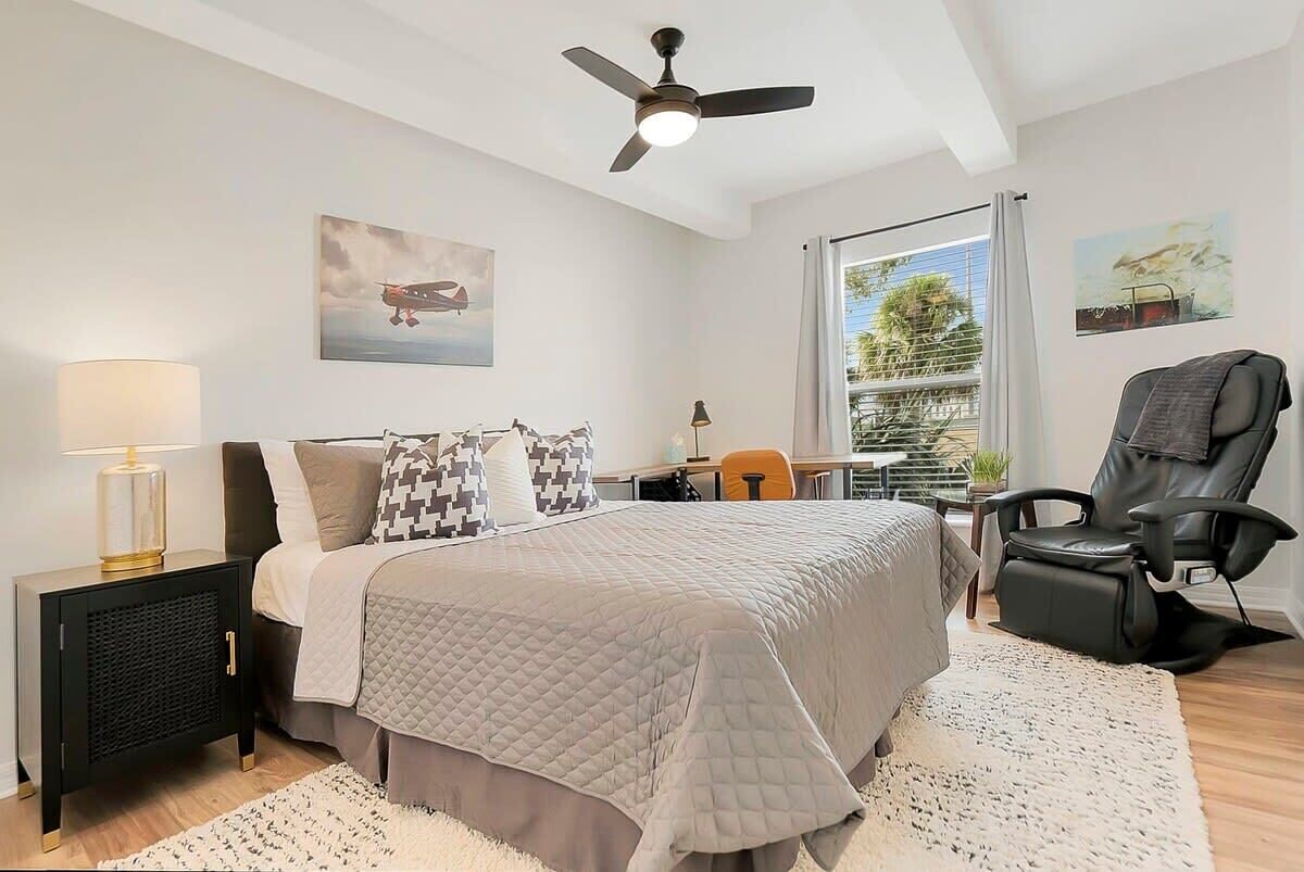 A bedroom with a bed, a desk, a chair, a ceiling fan, and a window with a view of greenery.