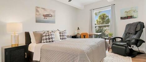 A bedroom with a bed, a desk, a chair, a ceiling fan, and a window with a view of greenery.