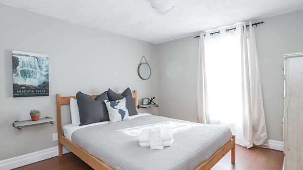 A bedroom with a wooden bed, grey bedding, a world map pillow, a small plant, and a poster of Niagara Falls. - Easy Walk to Clifton Hill Falls & Attractions! (Niagara Falls)