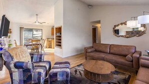 A living room with a brown leather sofa, patterned armchairs, and a coffee table. There is a dining area with a table and chairs, and a kitchen area with cabinets and a microwave.