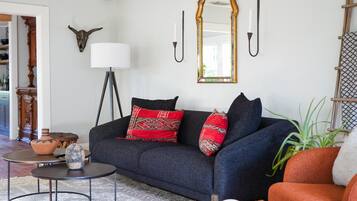 A living room with a dark blue sofa, red and patterned cushions, a small round coffee table, a floor lamp, a mirror, wall-mounted sconces, and a decorative bull skull.