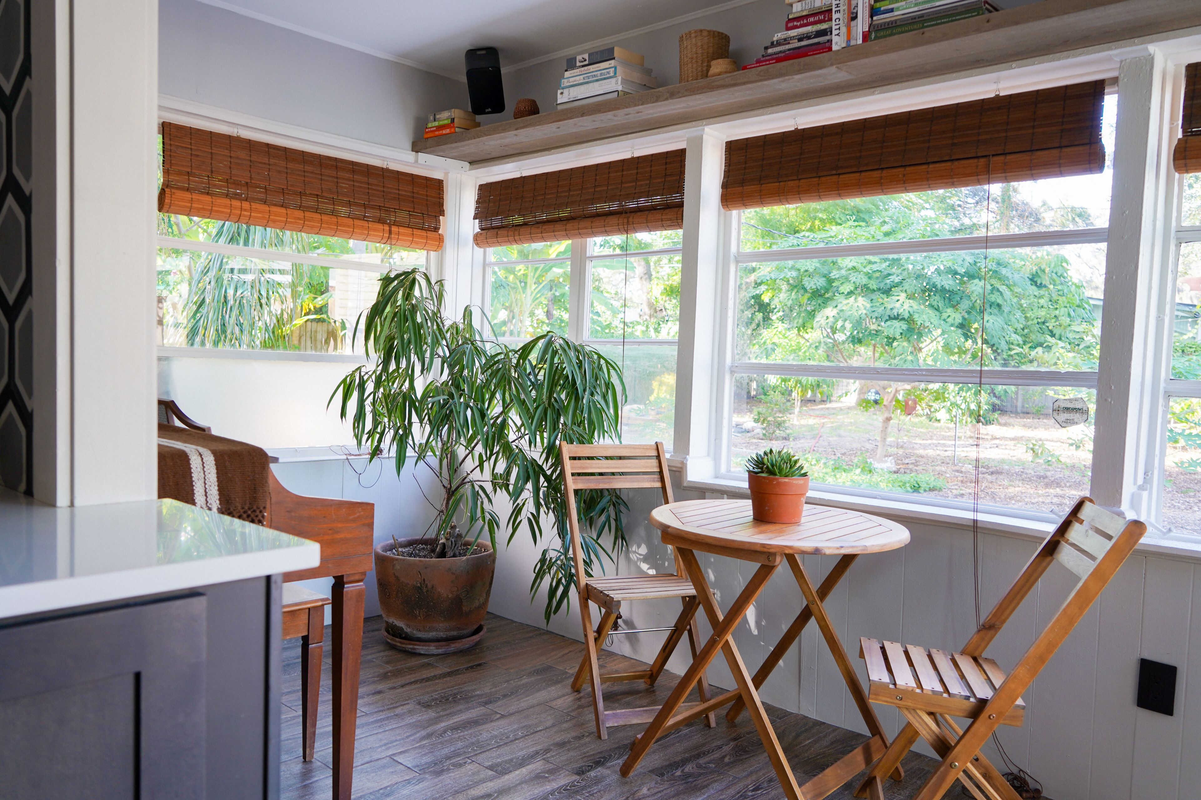 A sunroom with a wooden table and chairs, a potted plant, and large windows with blinds.