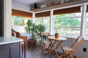 Una sala de estar con ventanales, una mesa y sillas de madera, una planta en maceta y persianas.