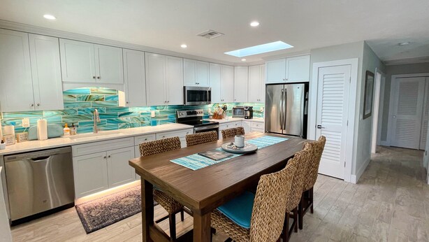 Una cocina moderna con armarios blancos, un refrigerador de acero inoxidable y un comedor con mesa de madera y sillas tejidas.
