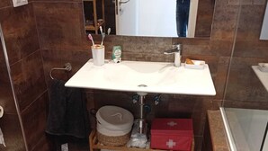 Et badeværelse med en hvid vask, et spejl og en bruseniche. Toiletartikler står på en hylde, og et håndklæde hænger på væggen.