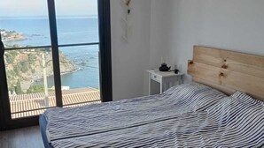 Et soveværelse med seng, et natbord og udsigt til havet og bygninger.