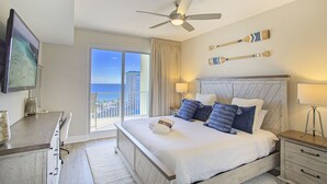 Une chambre d’hôtel avec un lit, un ventilateur de plafond, une télévision, un balcon avec vue sur l’océan et une tête de lit en bois.