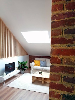 Ein moderner Loft-Raum mit geneigter Decke, einem weißen Sofa mit gelben und blauen Kissen, einem Holztisch, einem Topf mit Pflanze und einem Flachbildfernseher auf einem weißen Standfuß.