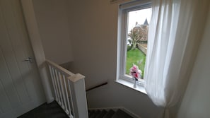 Un escalier avec une rampe blanche mène à un palier. Une fenêtre avec des rideaux blancs est visible et offre une vue sur un jardin et un bâtiment au loin.