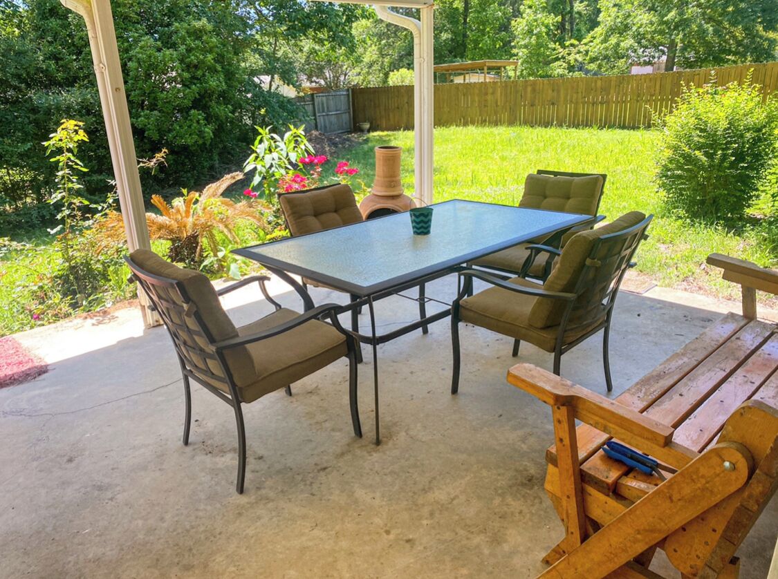 An outdoor patio with a table and four chairs, surrounded by greenery and a wooden fence.