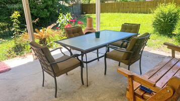 An outdoor patio with a table and four chairs, surrounded by greenery and a wooden fence.