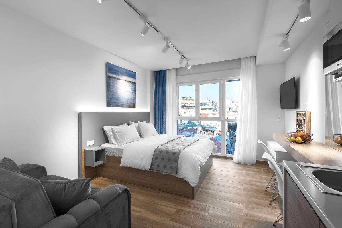 A modern hotel room with a large bed, a desk, and a view of the city through large windows.