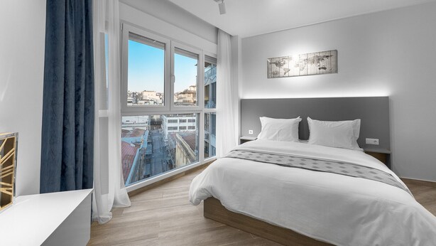 A modern hotel room with a large bed, a view of the city through floor-to-ceiling windows, and a minimalist design. - Hermes Nilie Hospitality MGMT (Thessaloniki)