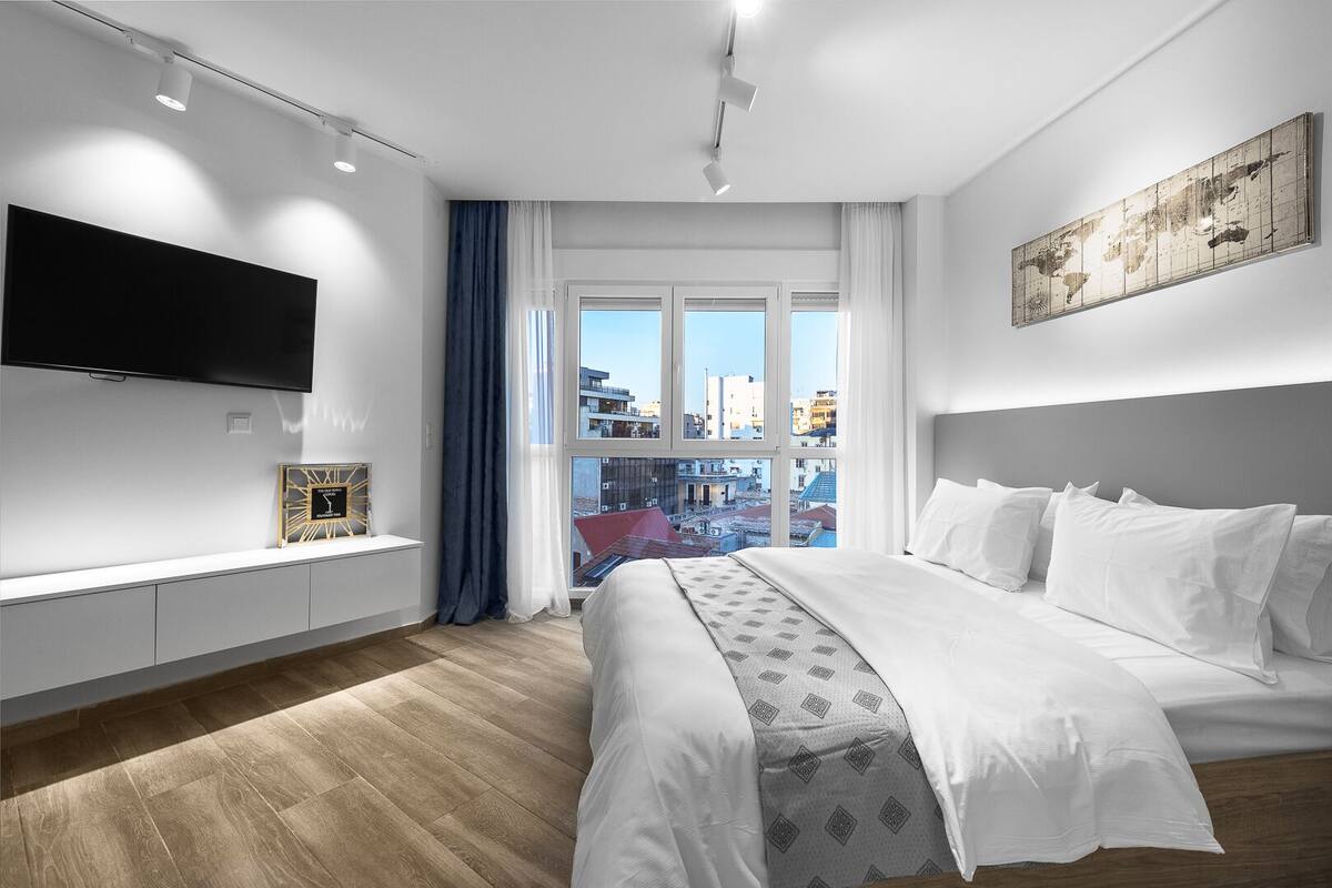 A modern hotel room with a large bed, a flat-screen TV mounted on the wall, a window with a city view, and a decorative world map on the wall.