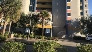 Un hôtel à plusieurs étages au design moderne, doté de balcons à chaque niveau, d’une cour intérieure agrémentée de palmiers et offrant une vue sur un ciel d’un bleu éclatant.