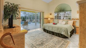 A bedroom with a bed, a desk, a television, a plant, and a view of the outdoors. - The Yellow | Lakefront Home, Heated Pool & Spa (Leigh Acres)